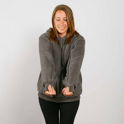 Woman wearing a gray sherpa zip hoodie with sherpa lining over a dark shirt, smiling with hands in pockets.