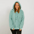 Woman wearing a seafoam color sherpa zip hoodie with hood up, standing against white background.