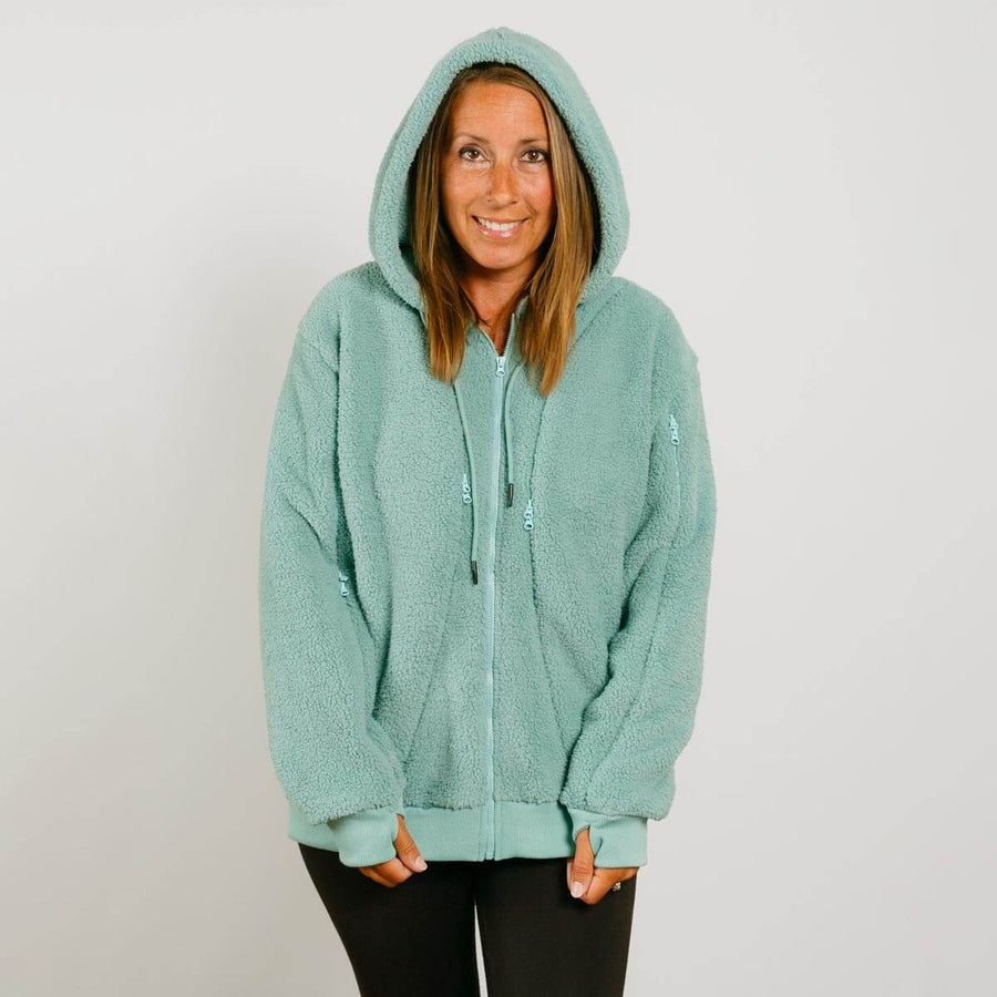 Woman wearing a seafoam color sherpa zip hoodie with hood up, standing against white background.