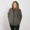Woman wearing a gray sherpa zip hoodie with hood up and hands in pockets, smiling at camera.