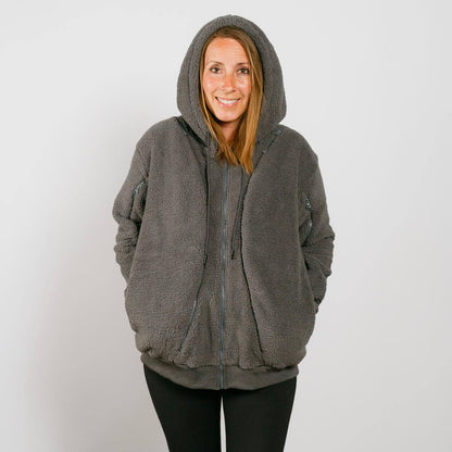 Woman wearing a gray sherpa zip hoodie with hood up and hands in pockets, smiling at camera.