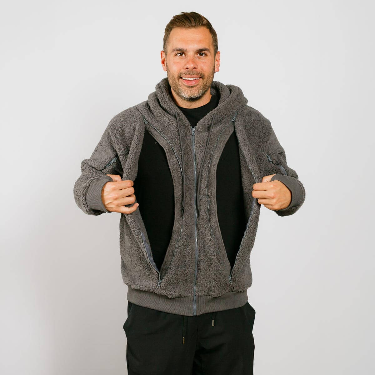 Man wearing gray sherpa zip hoodie over black shirt, holding jacket open against white background.
