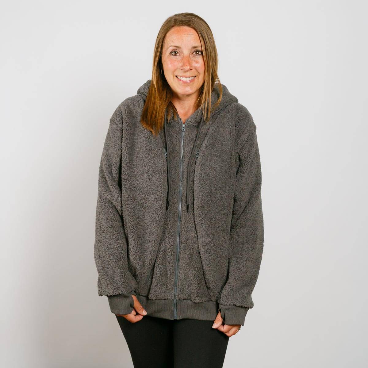 Woman wearing a gray sherpa zip hoodie with hood and black pants, smiling at camera.