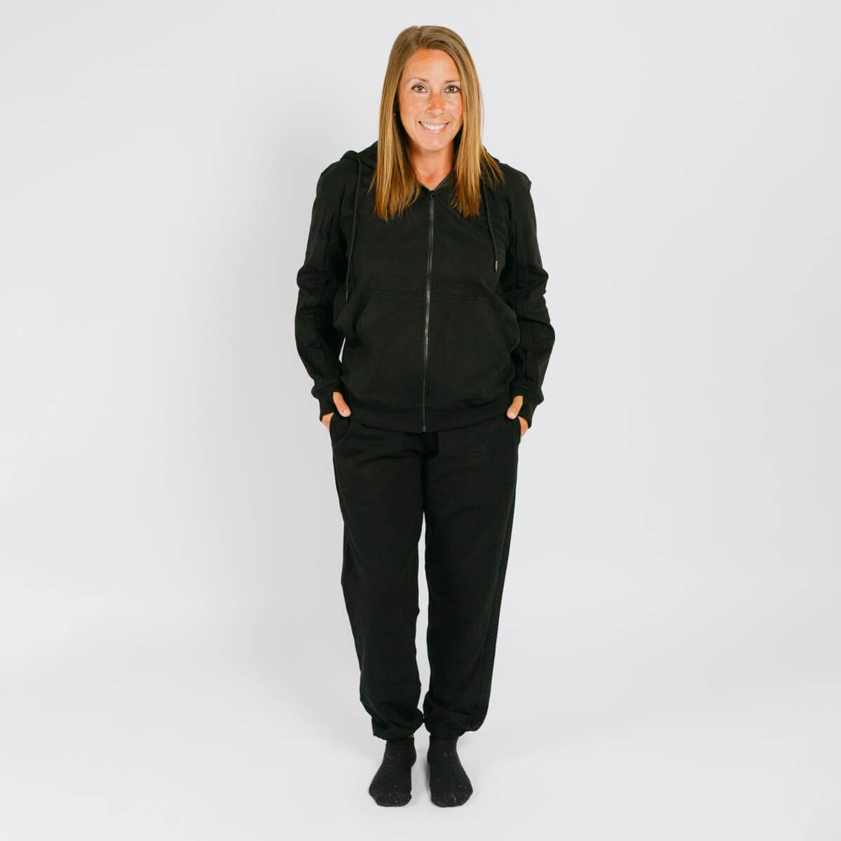 Woman wearing black zip-up hoodie and matching black sweatpants standing against white background. | Model is 5'3" 130lbs wearing size S