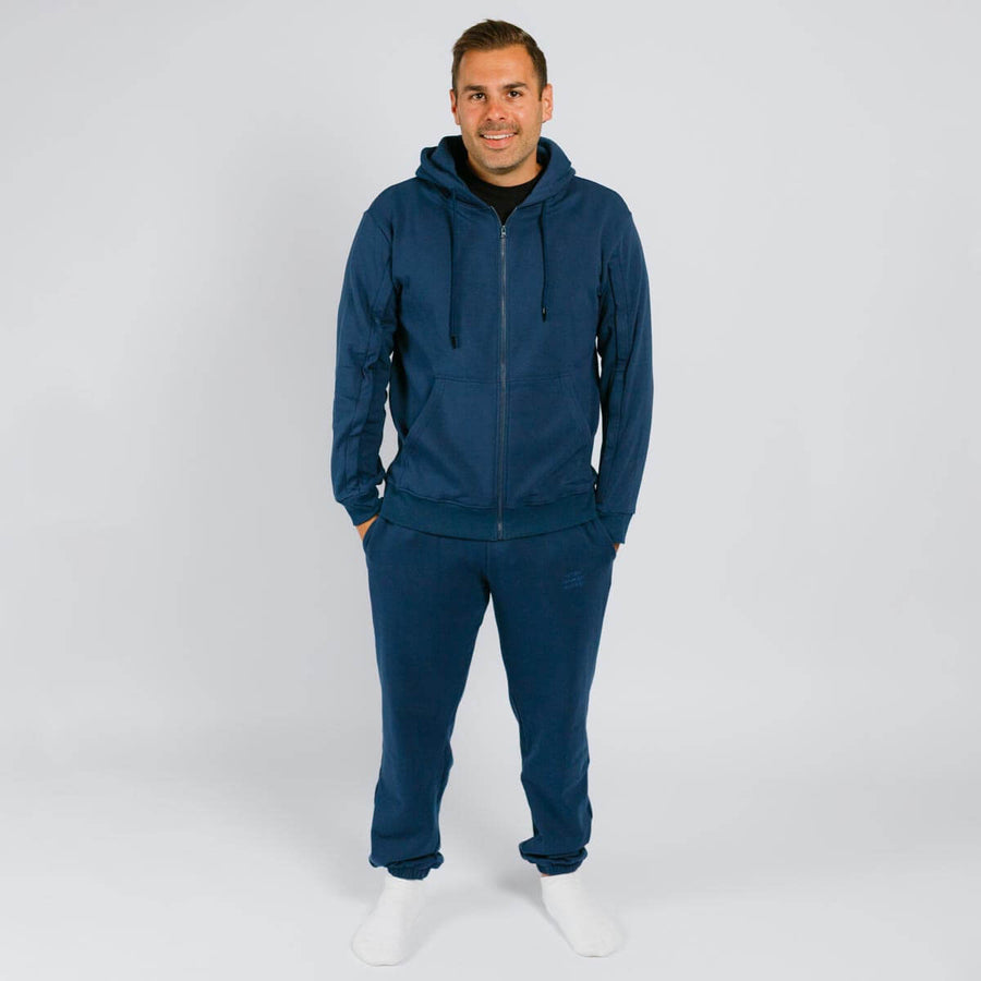 Man wearing dark blue zip-up hoodie and matching sweatpants standing against white background.