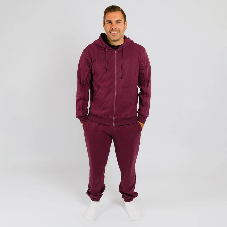Man wearing burgundy zip-up hoodie and matching sweatpants standing against white background.