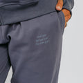 Person wearing slate blue sweatpants with embroidered text on the thigh, hands in pockets.