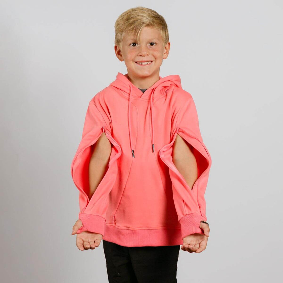 Young child wearing a coral pink hoodie with cutout sleeves and black pants, smiling at camera.